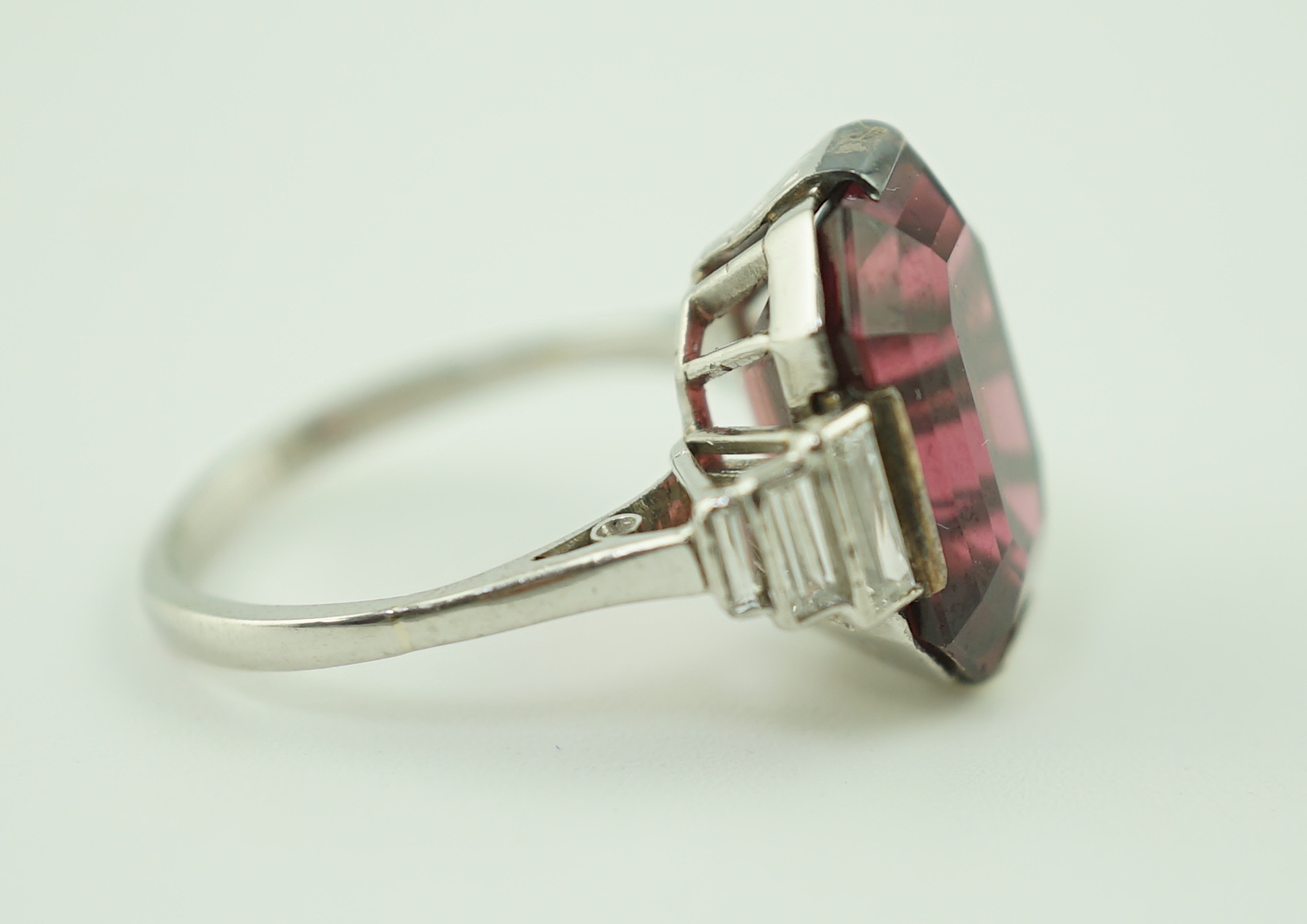 An Art Deco platinum and single stone emerald cut deep pink tourmaline set dress ring, with graduated six stone baguette cut diamond set shoulders